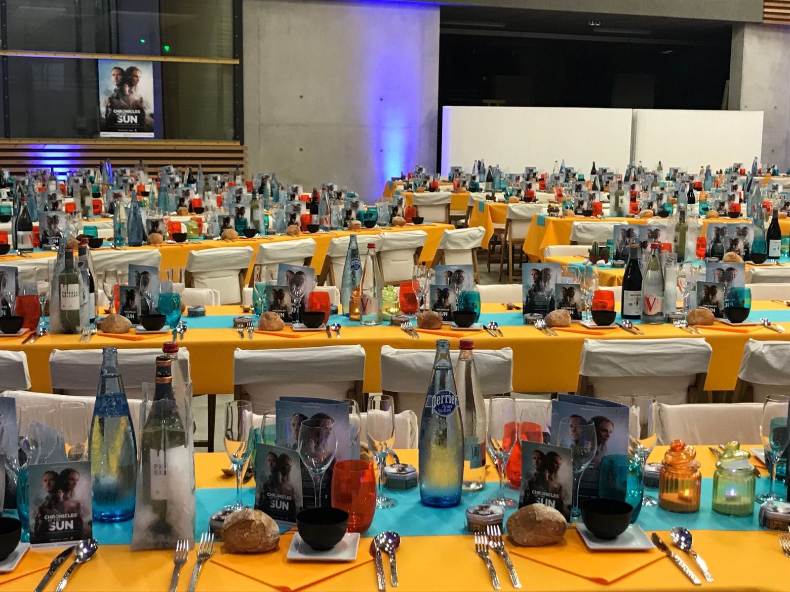 Salle du dîner de gala dressée en longues tables : nappes orange, chemins de table turquoise, menus et cartes de la série sur chaque couvert.