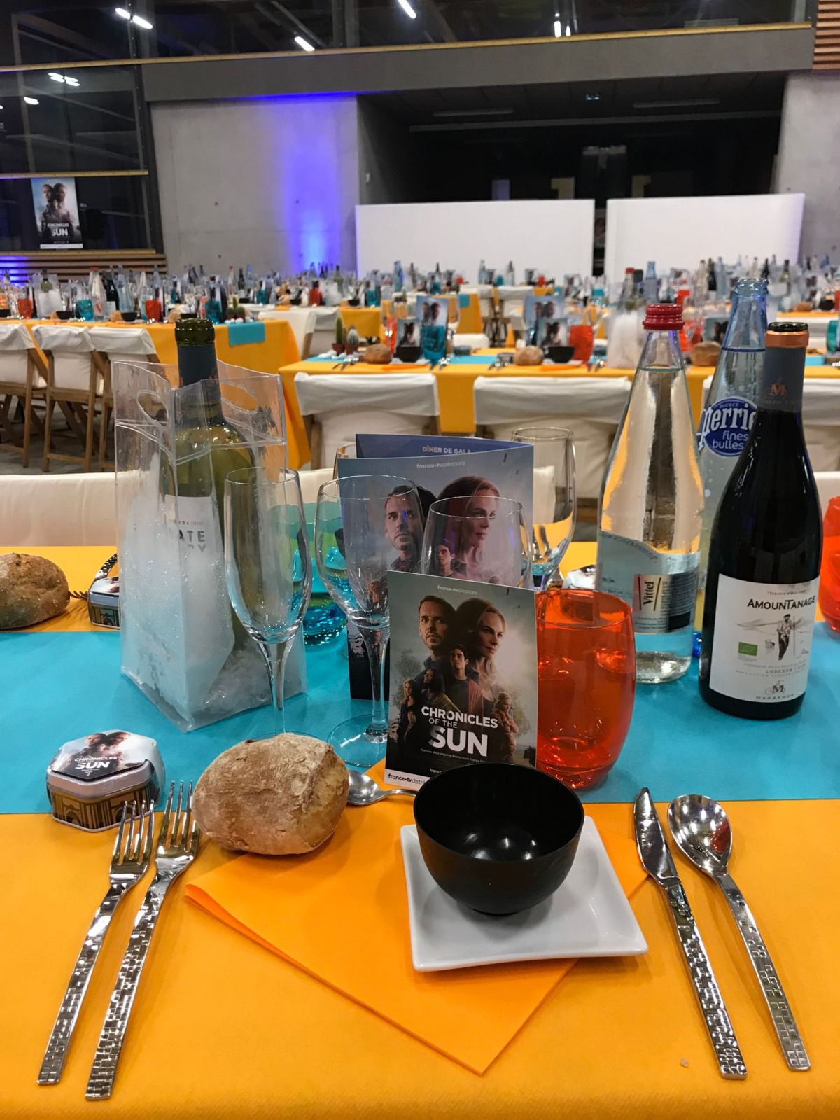 Couvert du dîner de gala : nappe orange, chemin de table turquoise, carte « Chronicles of the Sun », verres colorés et boîte hexagonale.