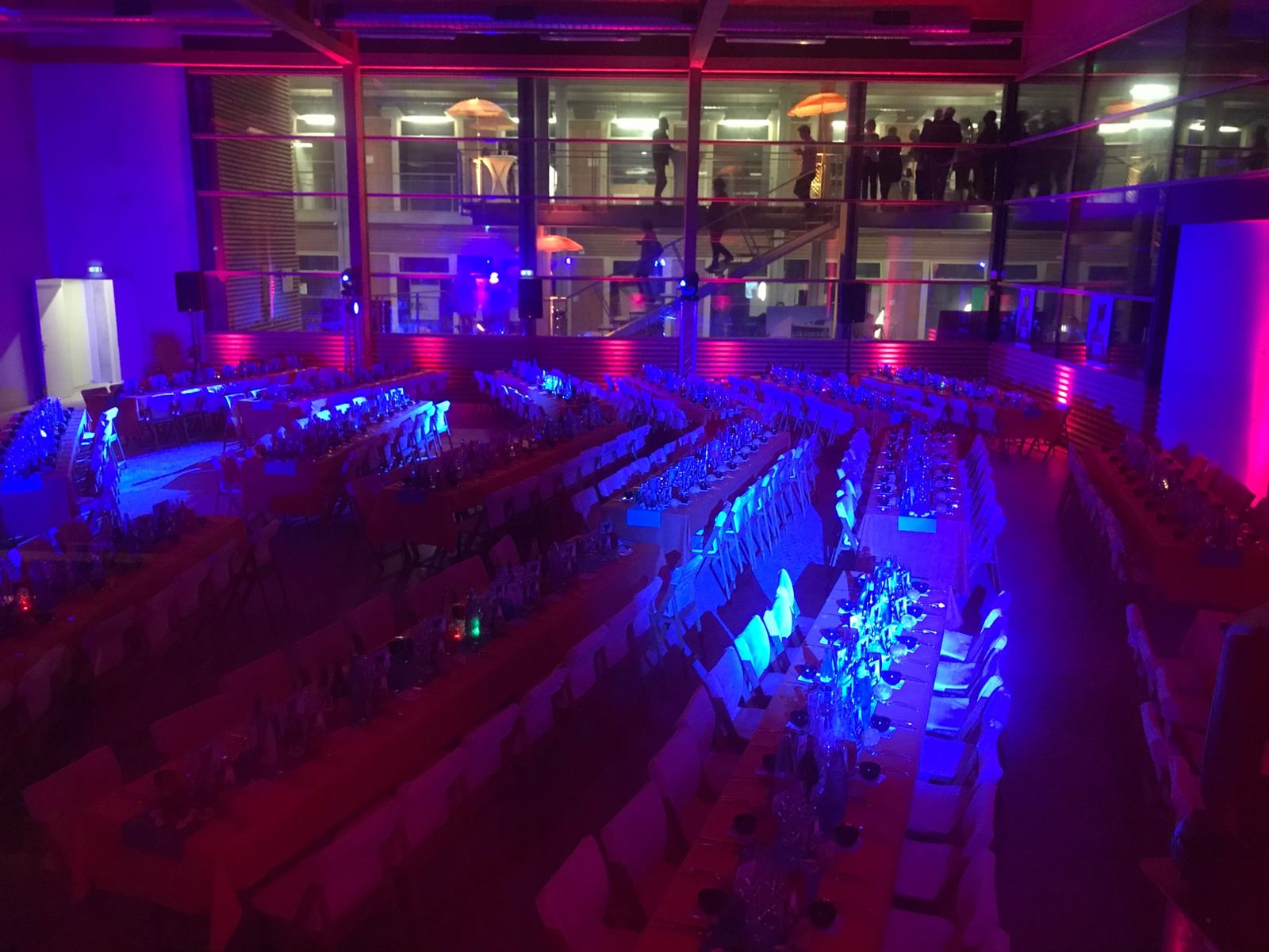 Vue plongeante sur la salle de gala en lumières bleues et roses, tables en vagues et mezzanine avec parasols.