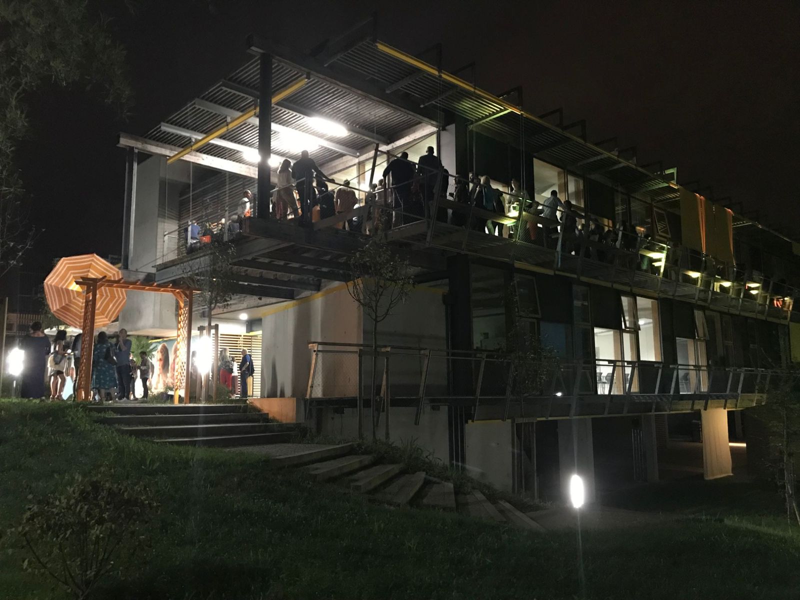 Terrasse du bâtiment illuminée de nuit : invités sur la coursive et parasol orange sous une pergola en bois.
