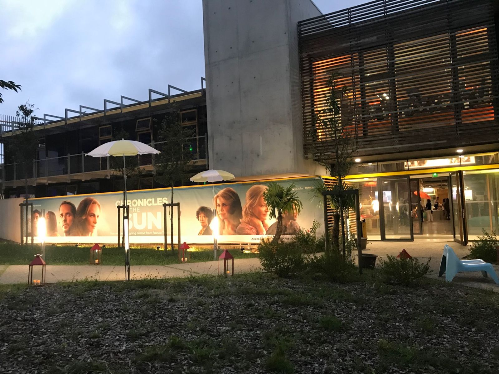 Espace Arkinova au crépuscule : banderole de 35 mètres de la série Chronicles of the Sun, parasols blancs et jaunes, palmier et lanternes rouges devant la façade en bois.